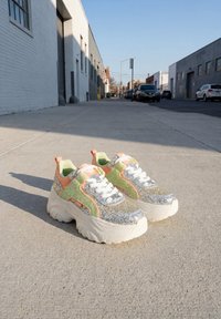 Kraftige sneakers med glitrende sølv, grønne og orange accenter placeret på en solbeskinnet fortov i et bymiljø.
