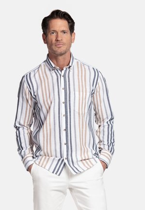 Man draagt een overhemd met lange mouwen, met verticale beige, grijze en witte strepen, gecombineerd met witte broek, handen in de zakken.