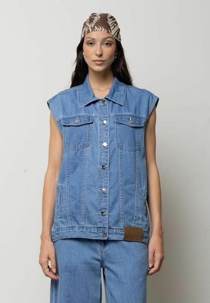 Donna con giacca di jeans blu senza maniche e pantaloni coordinati con foulard marrone fantasia in testa, in piedi davanti a uno sfondo chiaro e uniforme.