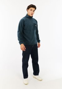 Sudadera de forro polar en verde azulado oscuro con cremallera de cuarto, que presenta un bolsillo en el pecho y puños acanalados. Combinada con jeans oscuros y zapatillas blancas.