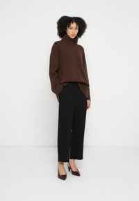 Suéter marrón de cuello alto tejido con detalles acanalados, combinado con pantalones negros de pierna ancha y tacones altos burdeos, sobre un fondo blanco.