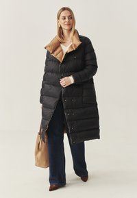 Parka nera con colletto marrone a contrasto, dotata di chiusura con bottoni, texture trapuntata e design fino al ginocchio. Indossata su jeans.