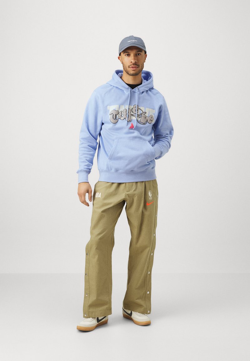 Sudadera azul claro con letras gráficas, pantalones de pinza beige con el logo de la NBA, y zapatillas blancas con acentos negros. Una gorra gris completa el look.