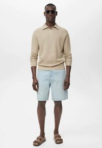 Suéter de punto beige con cuello con cremallera, pantalones cortos de mezclilla azul claro y sandalias de cuero marrón. Modelo usando gafas de sol, de pie contra un fondo blanco.