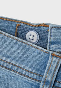 Jeans de denim de color azul claro con cierre de botón de plástico, costuras naranjas y una cinturilla texturizada. Primer plano del detalle de la cinturilla.