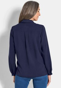 Marineblaue Langarmbluse mit Faltendetail am Rücken, geknöpften Manschetten und glatter Textur, kombiniert mit blauen Jeans.