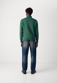 Sweatshirt vert avec un col montant, des poignets côtelés et une coupe décontractée associé à un jean droit bleu clair et des chaussures noires.