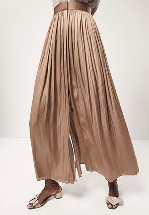 Brune plisserede bukser med brede ben kombineret med beige åbne, lave hæle med store sløjfer på mørkhudet model, der står.