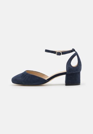 Chaussure à talon bloc bas en daim bleu marine avec bout rond fermé, détail découpé au talon et bride de cheville réglable avec boucle argentée.