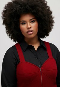 Red wool blend dress with a structured bodice and zip front, worn over a black button-up shirt. Features wide shoulder straps.