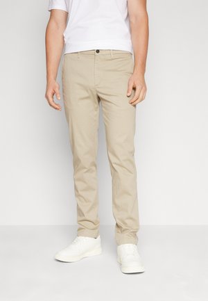 Mann trägt beige gerade geschnittene Hose, weißes Kurzarmhemd und weiße Turnschuhe, steht vor einem schlichten, hellen Hintergrund.