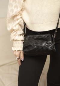 Borsa a tracolla intrecciata nera con texture liscia, forma rettangolare e una lunga tracolla, indossata sopra un maglione color crema chiaro e pantaloni neri.