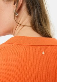 Pull orange texturé avec un col à côtes, comportant un petit charme en or à l'arrière du cou. Cheveux partiellement visibles.
