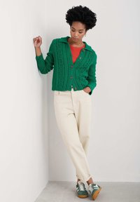 Grön stickad kofta med flätmönster och bruna knappar, över en röd topp, kombinerad med krämfärgade byxor och sneakers med gröna accenter.