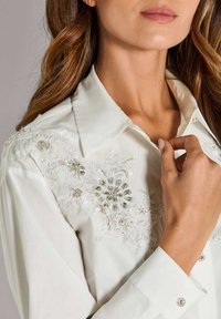 Witte blouse met bloemborduursel en kanten accenten, een gestructureerde kraag en knopen aan de mouwen, met nadruk op delicate texturen en details.