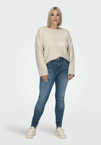 Pull crème à manches longues et en tissu texturé, associé à un jean bleu ajusté et des baskets de couleur claire. Tenue simple et décontractée.