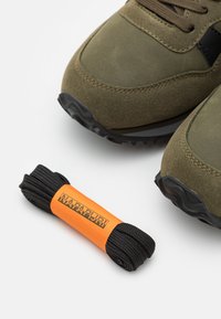 Oliwkowe sneakersy z zamszowym i siatkowym wierzchem, czarnymi akcentami i teksturowaną gumową podeszwą, w towarzystwie czarnych sznurowadeł w pomarańczowym opakowaniu.