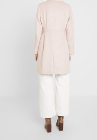 Manteau rose clair en mélange de laine à coupe droite, manches longues et détails de coutures au dos, associé à un pantalon blanc à jambes larges et des talons blocs embossés effet peau de serpent.