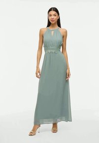 Mulher com cabelo escuro e comprido, vestindo um vestido verde-sálvia sem mangas e até ao chão, com faixa de renda na cintura e sapatos de salto dourados, de pé contra um fundo branco.