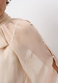 Durchsichtige beigefarbene Bluse mit hohem Kragen, sanften Drapierungen und Knopfdetails an den Ärmeln, die ein elegantes und leichtes Design zeigt.