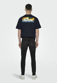Mørkeblå bomulls T-skjorte med et fargerikt grafisk motiv på ryggen, brukt med svarte jeans og hvite joggesko, sett fra baksiden.