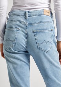 Jeans de denim azul claro con bolsillos traseros que presentan un detalle de costura en patrón de onda y una etiqueta de cuero marrón en la cinturilla.