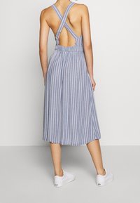 Jumpsuit rayé mi-long avec un design croisé dans le dos, une taille élastique et une couleur bleu clair avec des rayures blanches. Porté avec des baskets blanches.