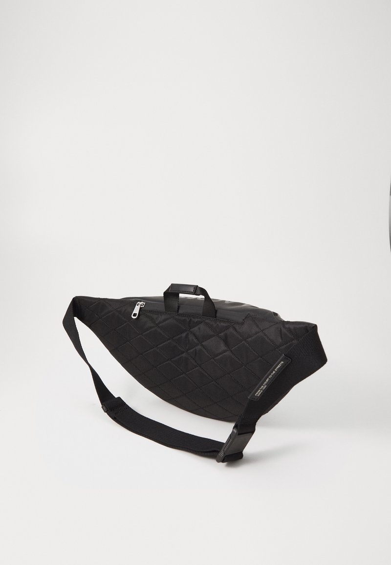 Sac en tissu matelassé noir avec une forme arrondie, doté d'une fermeture zippée, d'une poignée sur le dessus et d'une bandoulière amovible pour un transport facile.