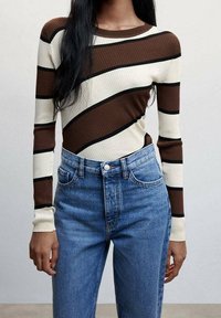 Nainen, joka pukeutuu ruskeaan, kermaan ja mustaan diagonaaliraidalliseen pitkähihaiseen paitaan ja korkeavyötäröisiin sinisiin farkkuihin.