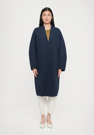 Navy blue long coat with a deep V-neckline, two front pockets, and oversized sleeves, paired with white pants and pointed white shoes.