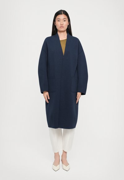 Abrigo largo azul marino con mangas anchas, bolsillos delanteros y escote en V profundo. Se lleva sobre una blusa oliva y pantalones blancos, combinado con zapatos puntiagudos color crema.