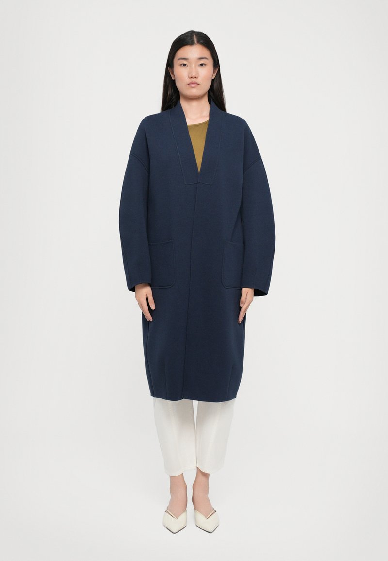 Manteau long bleu marine avec un décolleté en V profond, deux poches avant et des manches ballon, associé à un pantalon blanc et des chaussures blanches à talons pointus.