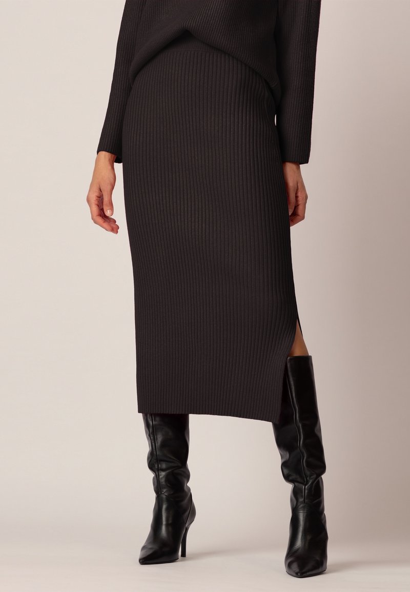 Femme portant une jupe midi côtelée noire avec une fente latérale et des bottes en cuir noir à talons hauts, debout devant un fond uni.