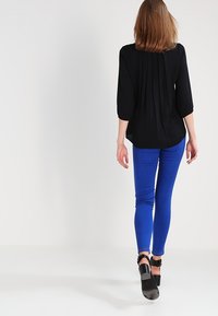 Svart blus med veck bak, kombinerad med åtsittande blå jeans. Outfitten kompletteras med svarta sandaletter med remmar och spänne vid vristen.