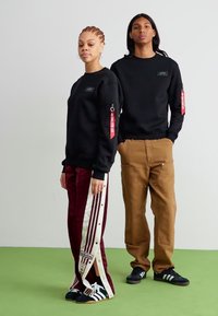 Zwarte sweatshirt met utility pocket, gecombineerd met kersenrode trainingsbroek met witte strepen. Beiden dragen zwarte sneakers. Groene vloer.