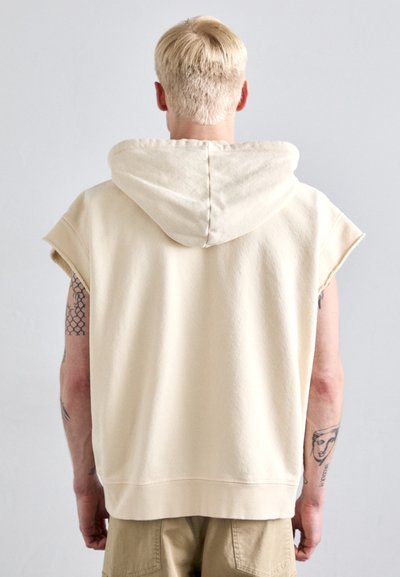Bež hoodie bez rukava s velikom kapuljačom i sirovim rubovima. Glatka majica od dukserice, opušteni kroj, vidljivi detalji šivanja na leđima.