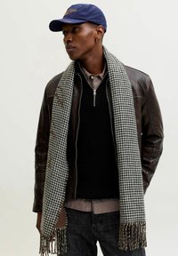 Halli ja musta hunditõllrisall, millel on narmad, kantud musta kootud kampsuni peal, mille all on ruuduline särk ja pruun nahkjakk.