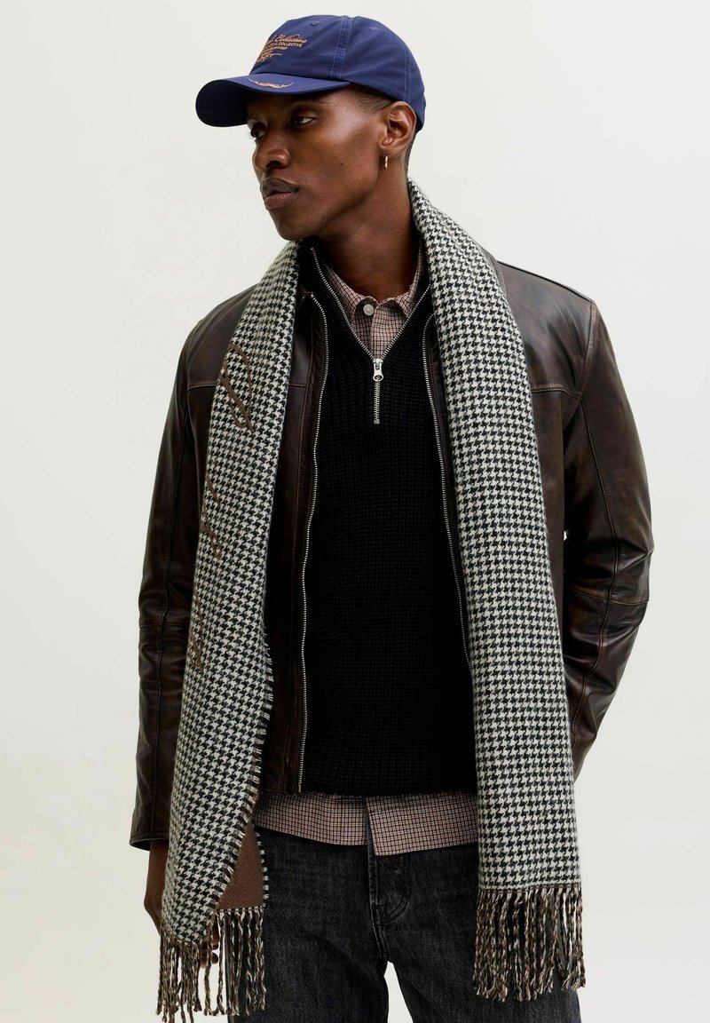 Halli ja musta hunditõllrisall, millel on narmad, kantud musta kootud kampsuni peal, mille all on ruuduline särk ja pruun nahkjakk.