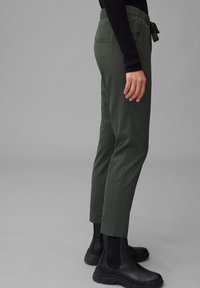 Person i olivgröna capribyxor med dragsko i midjan och svarta kraftiga Chelsea-boots mot en grå bakgrund.