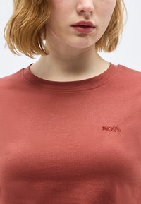 Άτομο με κοντά, ανοιχτό καστανά μαλλιά που φοράει πουκάμισο σε σκουριά χρώμα με μικρό κεντημένο λογότυπο "BOSS" στο στήθος.