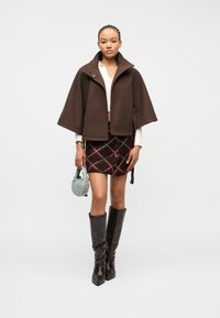 Νεαρή γυναίκα που φοράει καφέ κάπα, κοντή καρό φούστα, μαύρες μπότες μέχρι το γόνατο, κρατώντας μια μικρή τσάντα χειρός γαλάζιου χρώματος, στέκεται σε λευκό φόντο.