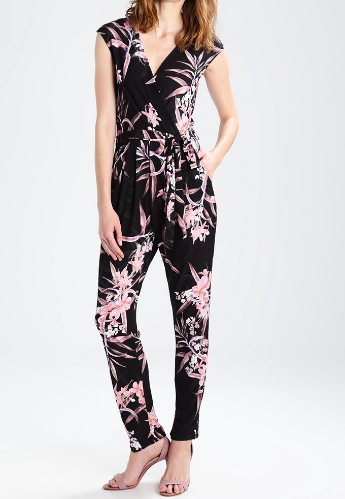 Femme portant une combinaison noire sans manches à fleurs roses et blanches et des talons ouverts couleur chair, debout avec une main dans la poche.
