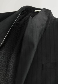 Schwarze gestreifte Anzugjacke mit strukturiertem Kragen. Das Innere verfügt über ein gemustertes Futter und eine einzelne Brusttasche. Subtile Designdetails durchgehend.