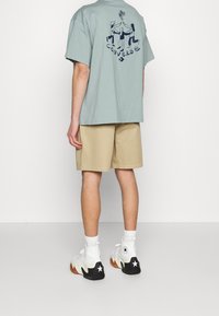 T-shirt di cotone verde chiaro con una grafica sulla schiena, abbinata a pantaloni corti beige e sneakers bianche con dettagli neri.