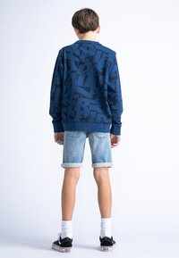 Felpa blu con motivo geometrico nero, abbinata a pantaloni corti in denim azzurro arrotolati e scarpe nere, vista da dietro.