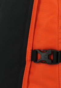 Sección de mochila de tela negra y naranja con superficie texturizada, que cuenta con una hebilla de plástico negro y costuras naranjas a lo largo de la costura.