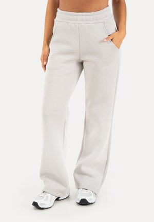 Pantalon de survêtement gris clair à jambes larges avec taille élastique et poches, porté avec des chaussures de sport blanches, mains dans les poches.
