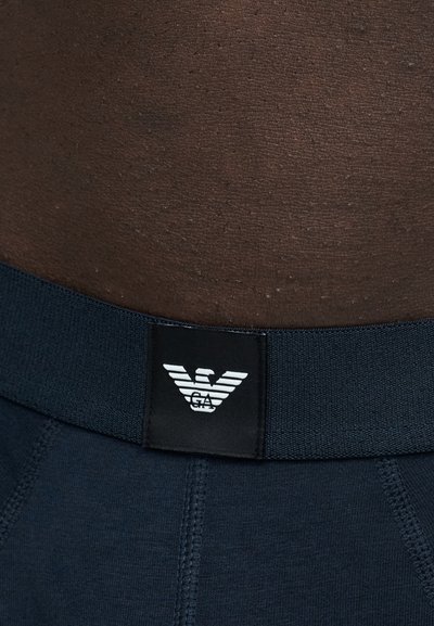 Gros plan d'une taille à la peau foncée portant un sous-vêtement bleu marine avec une ceinture noire comportant un logo d'aigle blanc.