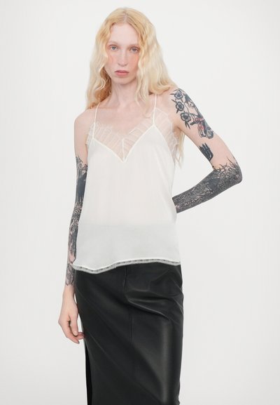 Camisole alb cu bretele subțiri și decolteu în V, având detalii din dantelă pe umeri și un finisaj neted, în straturi. Asortat cu o fustă neagră.