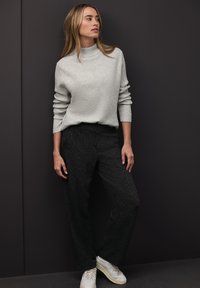 Hellgrauer, gerippter Pullover mit hohem Kragen, kombiniert mit dunkel strukturierten Hosen und weißen Turnschuhen, vor einem dunklen Hintergrund.